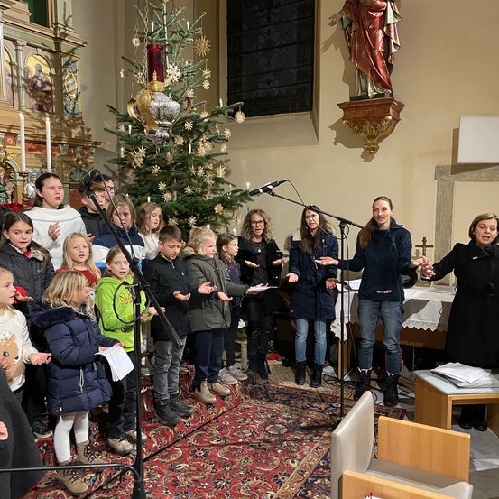 Ein Kinderchor steht in einer Kirche, singt und klatscht in die Hände. Hinter ihnen steht ein Weihnachtsbaum, der mit Lichtern und Ornamenten geschmückt ist. Rechts ist eine Statue eines Heiligen an der Wand angebracht.