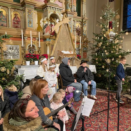 Eine Gruppe von Menschen versammelt sich in einer Kirche für eine Aufführung. Einige sitzen, andere stehen, darunter ein Junge mit einem Mikrofon. Musikinstrumente wie Gitarre und Klavier sind vorhanden. Weihnachtsdekorationen schmücken den Altar und den Hintergrund.