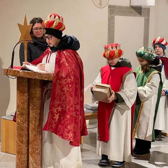 Eine Gruppe von Kindern in roten und weißen Gewändern und Kronen steht in einer Kirche, einer von ihnen liest ein Buch am Pult.