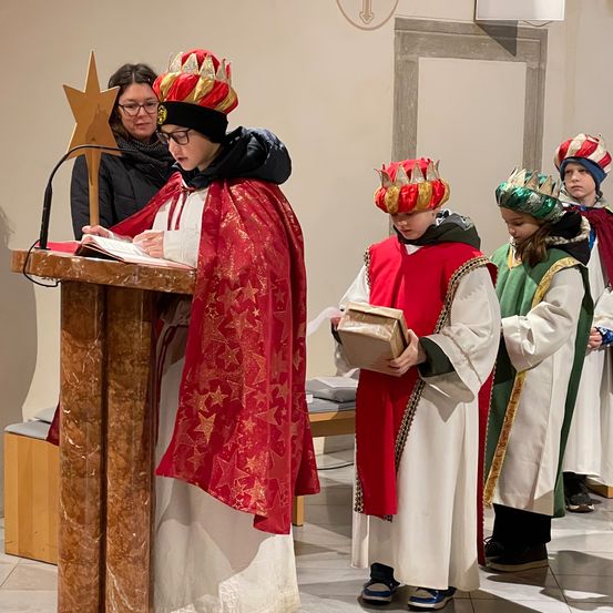 Eine Gruppe von Kindern in roten und weißen Gewändern und Kronen steht in einer Kirche, einer von ihnen liest ein Buch am Pult.