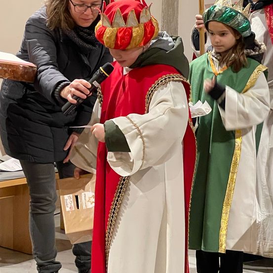 Eine Frau führt ein Kind in einem roten und weißen Kostüm mit goldener Krone, während sie ein Mikrofon hält. Ein anderes Kind in einem grünen Kostüm steht in der Nähe.