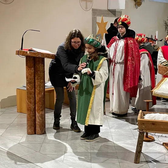 Eine Frau hilft einem Kind, das eine grüne Robe und Krone trägt, ein Mikrofon bei einer Kirchenveranstaltung zu halten.