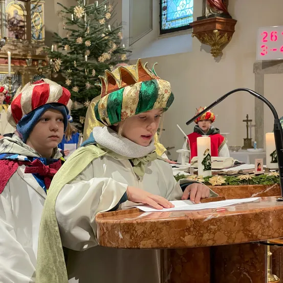 Kinder in festlichen Kostümen stehen vor einem Altar, eines liest von einem Papier, während ein anderes zusieht. Dahinter sind ein Weihnachtsbaum, Kerzen und eine Uhr zu sehen.