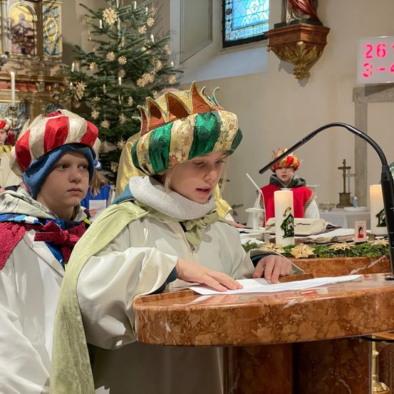 Kinder in festlichen Kostümen stehen vor einem Altar, eines liest von einem Papier, während ein anderes zusieht. Dahinter sind ein Weihnachtsbaum, Kerzen und eine Uhr zu sehen.