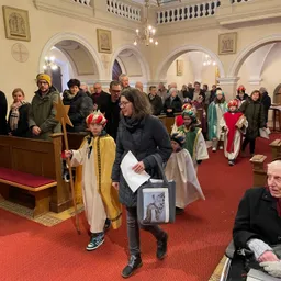 Eine Gruppe von Menschen geht in einer Kirche. Einige sind in Kostümen. Eine Frau hält ein Papier und eine Tasche. Ein Kind hält einen Stab. Andere beobachten.