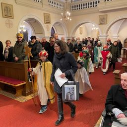 Eine Gruppe von Menschen geht in einer Kirche. Einige sind in Kostümen. Eine Frau hält ein Papier und eine Tasche. Ein Kind hält einen Stab. Andere beobachten.