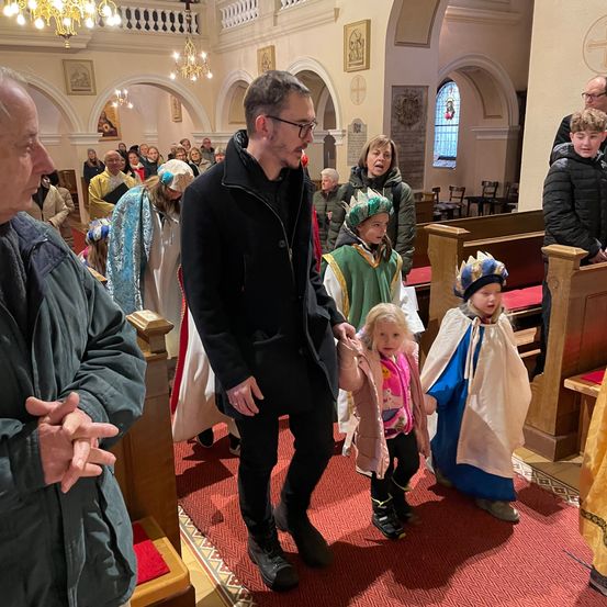Eine Gruppe von Menschen in einer Kirche, einige in Kostümen, ein Mann in einem schwarzen Mantel führt zwei Kinder in rosa und grünen Kostümen.