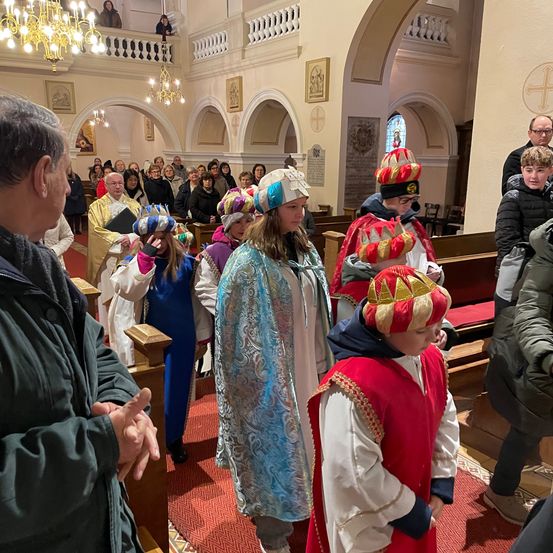 Eine Gruppe von Kindern in Kostümen steht in einem Kirchenschiff, einige tragen Hüte, mit Erwachsenen im Publikum.