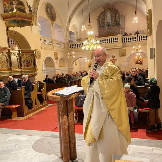 Ein Priester in goldener Robe spricht in ein Mikrofon in einer Kirche, während eine Menge auf den Kirchenbänken sitzt.