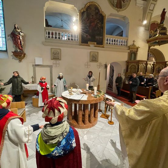 Eine religiöse Zeremonie in einer Kirche mit Menschen in traditionellen Kostümen und einem Priester, der den Gottesdienst leitet. Altar mit Kerzen und Papieren.
