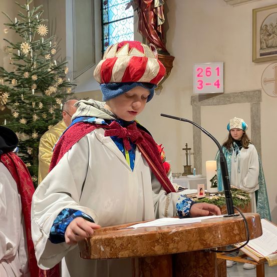 Eine Person in einem roten und weißen Gewand mit einem Hut steht an einem Rednerpult in einer Kirche. Hinter ihr steht ein geschmückter Weihnachtsbaum. An der Wand befindet sich ein Schild mit der Nummer 261.