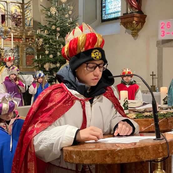 Ein Kind in einem roten und weißen Gewand mit Krone liest von einem Skript an einem Podium in einer Kirche. Hinter ihnen stehen und sitzen andere in ähnlicher Kleidung. Kerzen und ein Weihnachtsbaum sind im Hintergrund zu sehen.