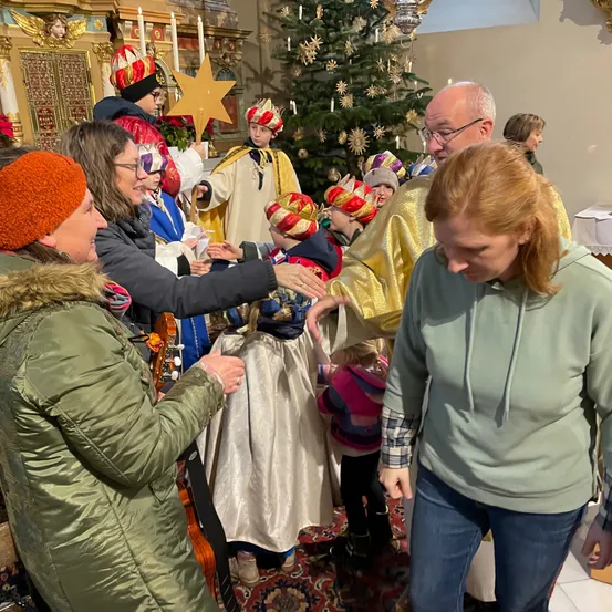 Eine Gruppe von Erwachsenen und Kindern in festlichen Kostümen versammelt sich um einen geschmückten Weihnachtsbaum in einer Kirche. Ein Mann in einem goldenen Gewand hält einen Stern, während eine Frau in einer grünen Jacke eine Geige hält.
