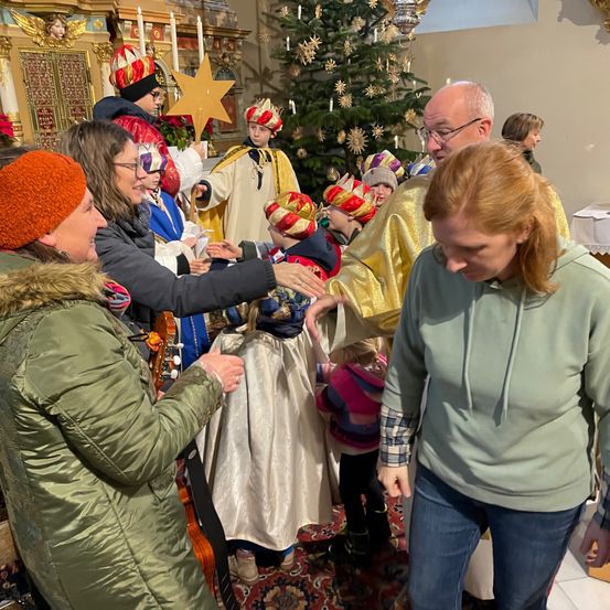 Eine Gruppe von Erwachsenen und Kindern in festlichen Kostümen versammelt sich um einen geschmückten Weihnachtsbaum in einer Kirche. Ein Mann in einem goldenen Gewand hält einen Stern, während eine Frau in einer grünen Jacke eine Geige hält.