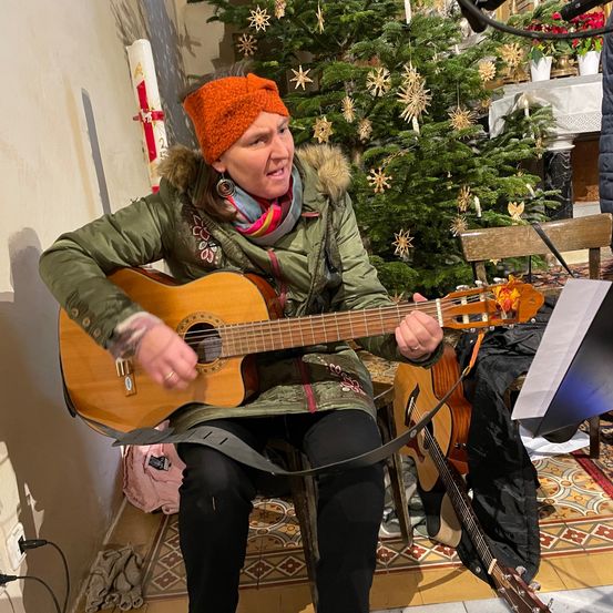 Eine Frau in einer grünen Jacke und einer orangefarbenen Kopfband spielt Gitarre, während sie vor einem Weihnachtsbaum sitzt.