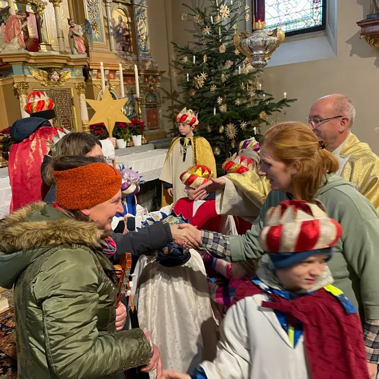 Eine Gruppe von Menschen in festlicher Kleidung, einschließlich Kinder in Kronen, versammelt sich in einer Kirche um einen Weihnachtsbaum. Sie schütteln Hände mit Erwachsenen, möglicherweise während einer Feierlichkeit.