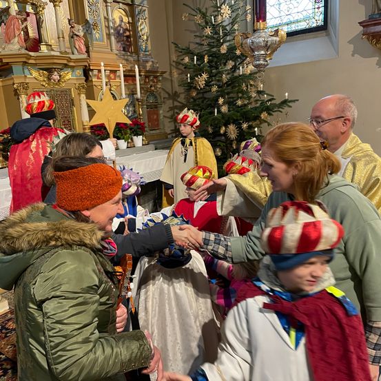 Eine Gruppe von Menschen in festlicher Kleidung, einschließlich Kinder in Kronen, versammelt sich in einer Kirche um einen Weihnachtsbaum. Sie schütteln Hände mit Erwachsenen, möglicherweise während einer Feierlichkeit.