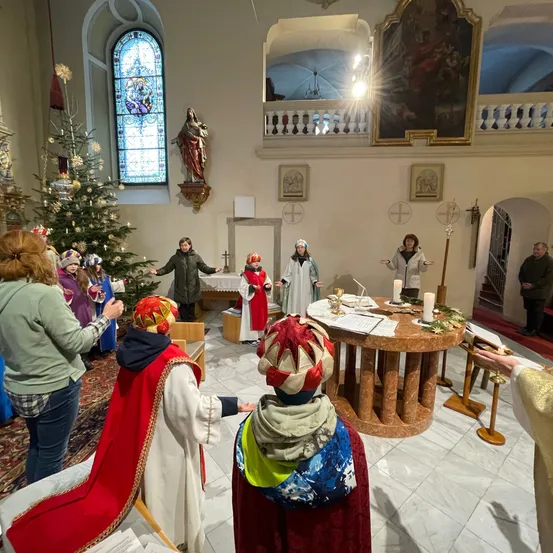 Eine Gruppe von Menschen in festlicher Kleidung versammelt sich in einer Kirche, mit einem Weihnachtsbaum und einer Statue im Hintergrund. Ein Tisch mit Kerzen und Papieren ist aufgestellt, und eine Person hält ein Mikrofon.