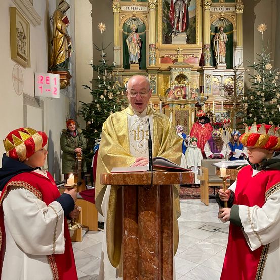 Ein Priester hält eine Predigt bei einer Kirchenzeremonie, wobei zwei Kinder Kerzen halten. Hinter ihm steht ein geschmückter Altar mit Weihnachtsbäumen und Statuen.