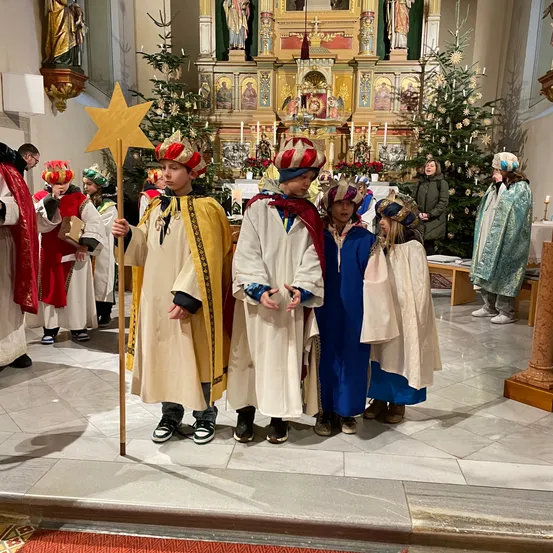 Eine Gruppe von Kindern in traditionellen Kostümen steht während einer Weihnachtszeremonie in einer Kirche, hält einen Stern und steht vor einem geschmückten Altar.