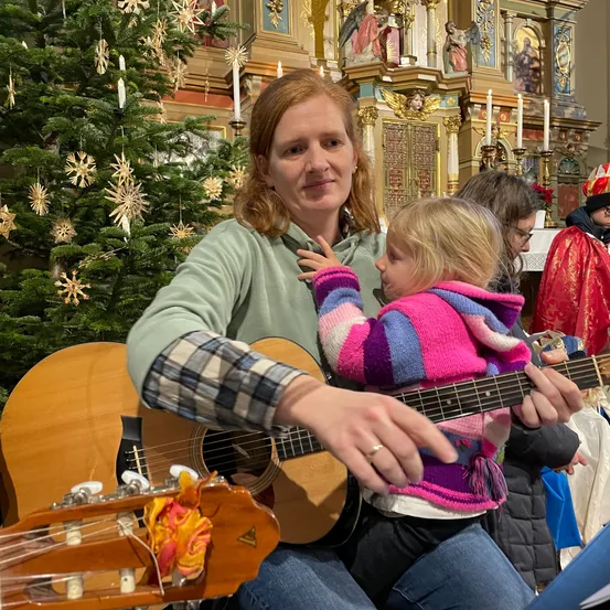 Eine Frau sitzt auf einer Bank mit einem kleinen Kind auf dem Schoß und spielt Gitarre in einer Kirche. Hinter ihnen steht ein mit Ornamenten geschmückter Weihnachtsbaum, und ein Priester steht in der Nähe.