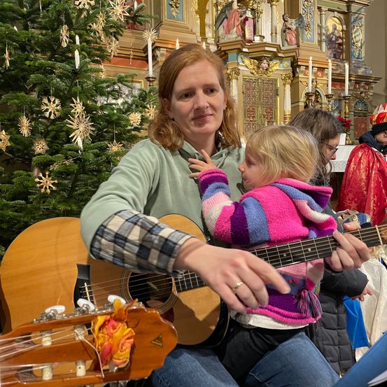 Eine Frau sitzt auf einer Bank mit einem kleinen Kind auf dem Schoß und spielt Gitarre in einer Kirche. Hinter ihnen steht ein mit Ornamenten geschmückter Weihnachtsbaum, und ein Priester steht in der Nähe.