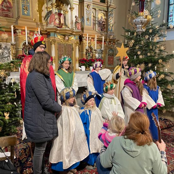 Eine Gruppe von Kindern in bunten Kostümen, möglicherweise bei einer Nachstellung der Weihnachtsgeschichte, steht in einer Kirche. Erwachsene und Kinder beobachten und lauschen. Ein Weihnachtsbaum und Kerzen sind im Hintergrund.