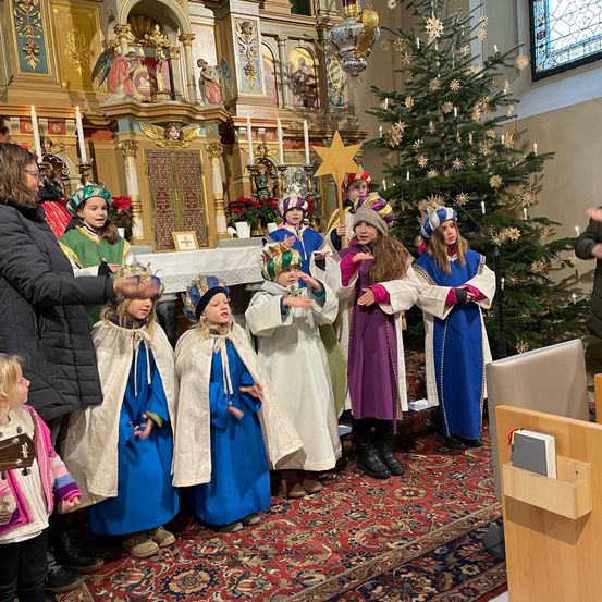 Eine Gruppe von Kindern in Kostümen und Mützen singt in einer Kirche mit einem Erwachsenen und einem Weihnachtsbaum.