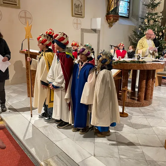Kinder in bunten Kostümen stehen in einer Kirche, halten einen Stern und ein Priester liest aus einem Buch.