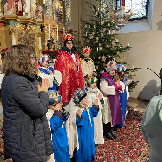 Mehrere Kinder in Kostümen singen in einer Kirche. Ein Mann in einem roten Umhang steht vor ihnen. Ein Weihnachtsbaum mit brennenden Kerzen steht in der Nähe.