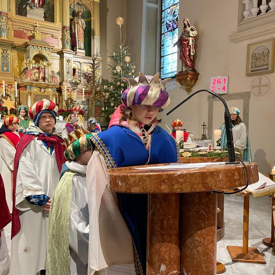 In einer Kirche stehen Kinder mit bunten Kronen um ein Podest mit Mikrofon. Hinter ihnen steht eine Statue von Jesus, und der Altar ist mit Kerzen und einem Weihnachtsbaum geschmückt.