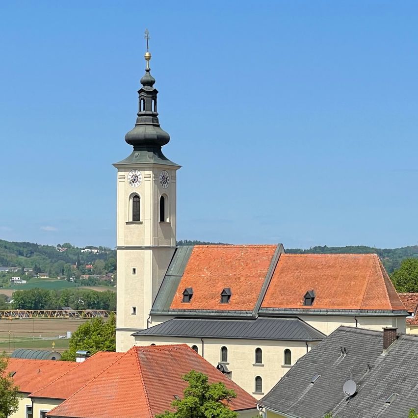 An aerial view of a church with a tower and a steeple, surrounded by a cluster of houses with orange roofs. The area is covered with lush green trees, and there are hills in the distance.