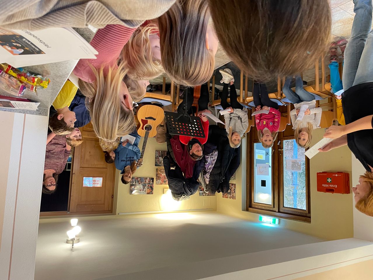 Eine Gruppe von Kindern steht in einem Klassenzimmer. Einige halten Gitarren und Musikblätter, während andere von Papieren lesen. Der Raum hat gelbe Wände, eine Holztür und ein Fenster mit Blick nach draußen.