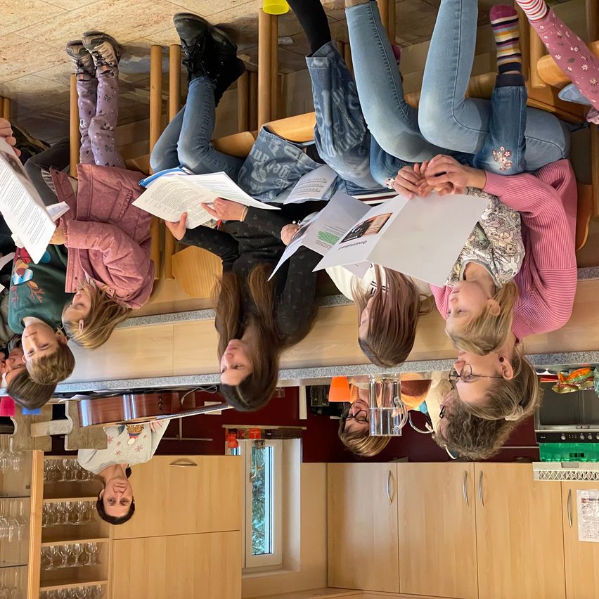 Eine Gruppe von Menschen versammelt sich in einem Raum, einige sitzen auf Stühlen und andere stehen. Sie halten Papiere in den Händen und scheinen etwas zu besprechen. Der Raum hat einen Holzschrank, ein Regal mit Weingläsern und ein Fenster. Eine Person in der Mitte hält eine Geige.