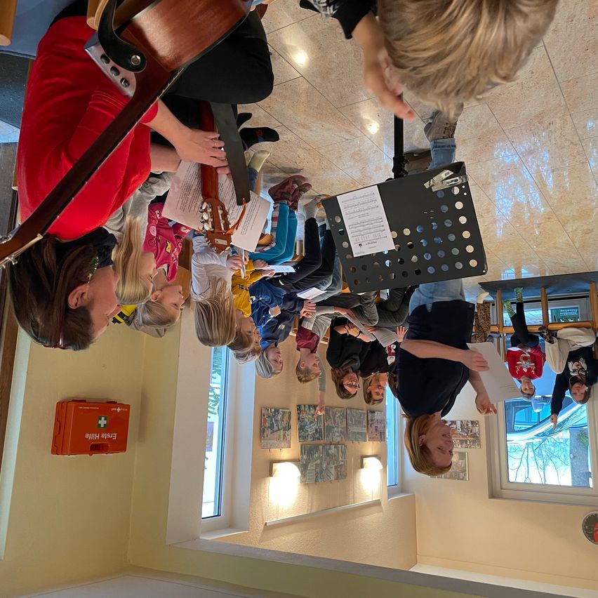 Eine Gruppe von Menschen versammelt sich in einem Raum und hält Gitarren und Papiere. Sie scheinen für eine Aufführung zu proben.