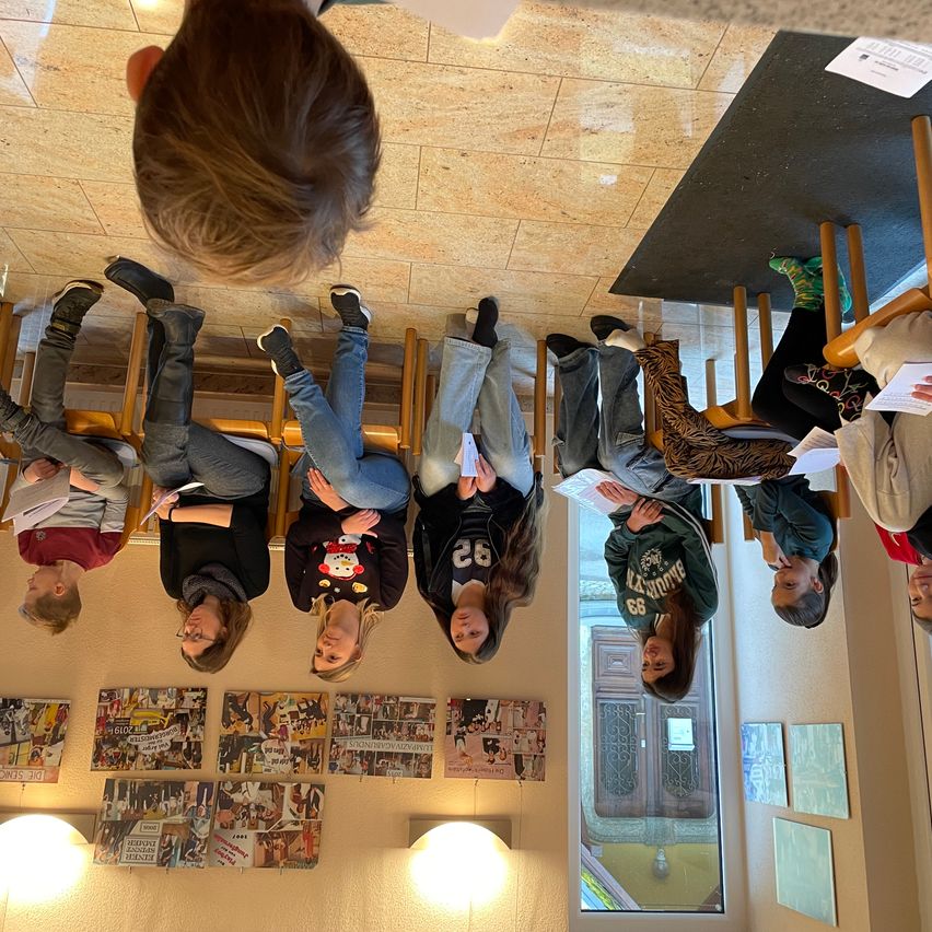 Eine Gruppe von Menschen steht kopfüber in einem Raum und hält Papiere. Sie bilden einen Kreis mit einer Tür und einem Fenster im Hintergrund.