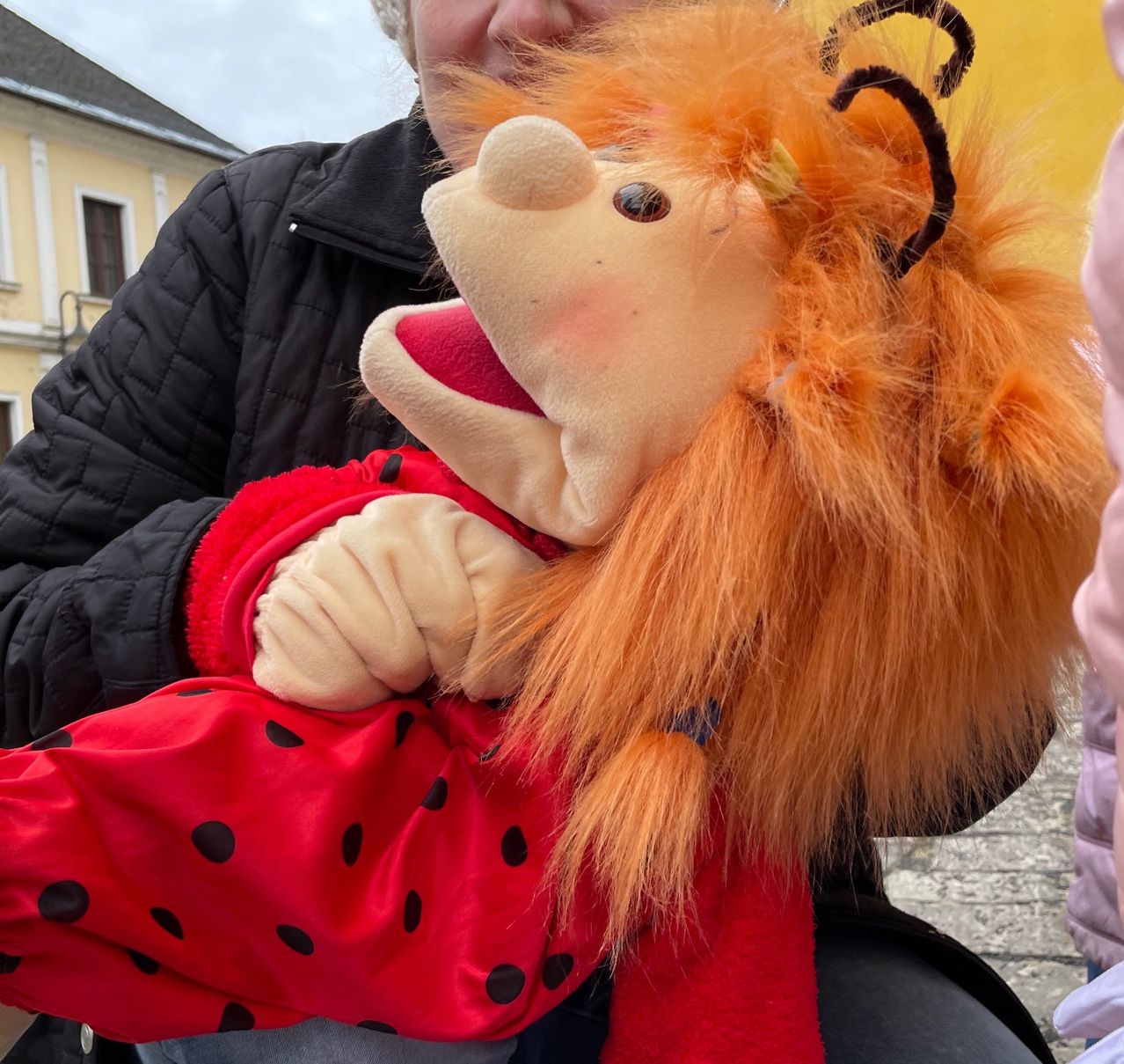 Eine Person hält eine Puppe in Rot mit schwarzen Punkten, die auf einer Kopfsteinpflasterstraße vor einem gelben Gebäude steht.