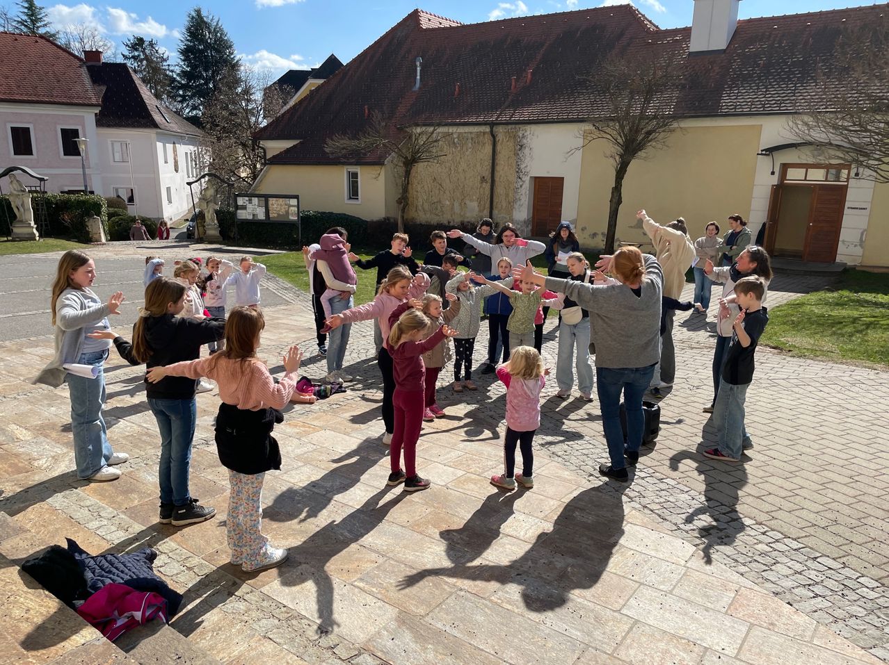 Eine Gruppe von Kindern und Erwachsenen tanzt an einem sonnigen Tag im Freien vor einem Gebäude. Einige sind barfuß.