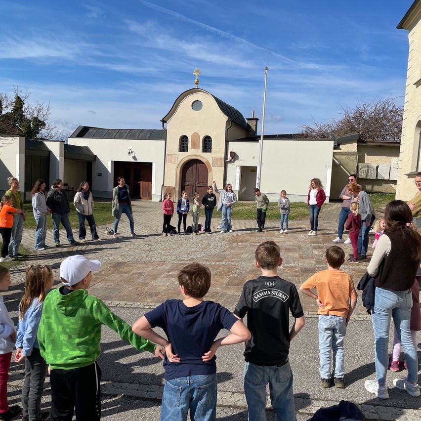Eine Gruppe von Menschen versammelt sich vor einer Kirche, einige stehen und andere schauen sich um. Sie tragen Freizeitkleidung und Turnschuhe. In der Mitte schauen sich zwei Personen etwas an.