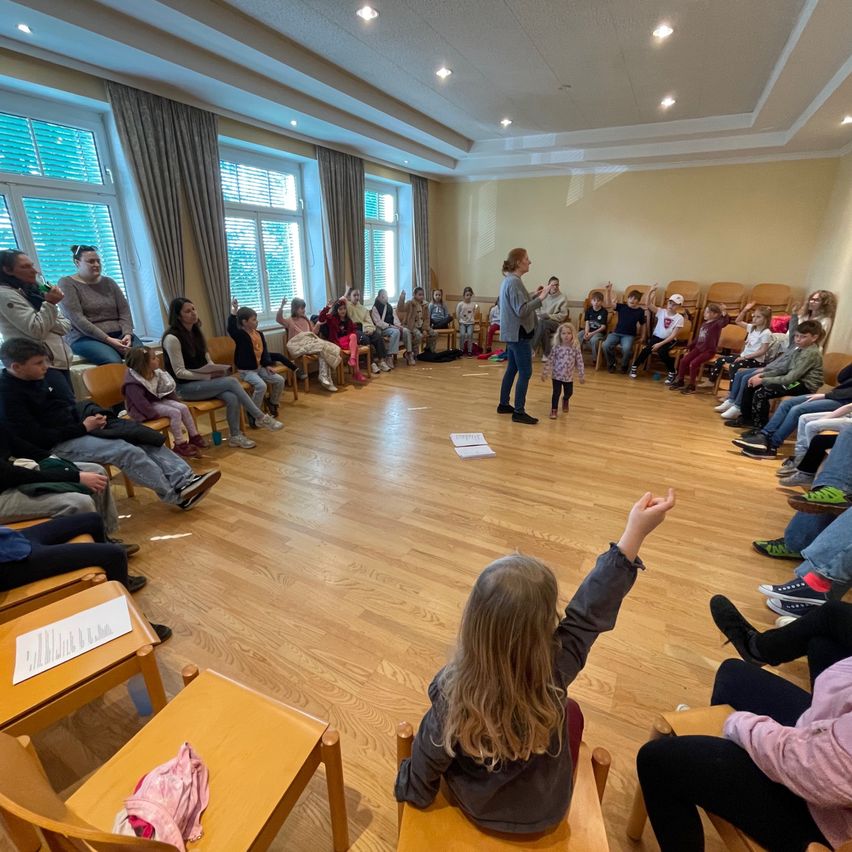 Eine Gruppe von Menschen sitzt in Stühlen um eine Frau und ein Kind in einem Raum mit Holzböden und großen Fenstern. Die Frau spricht und das Kind hört zu. Auf einigen Stühlen liegen Papiere.