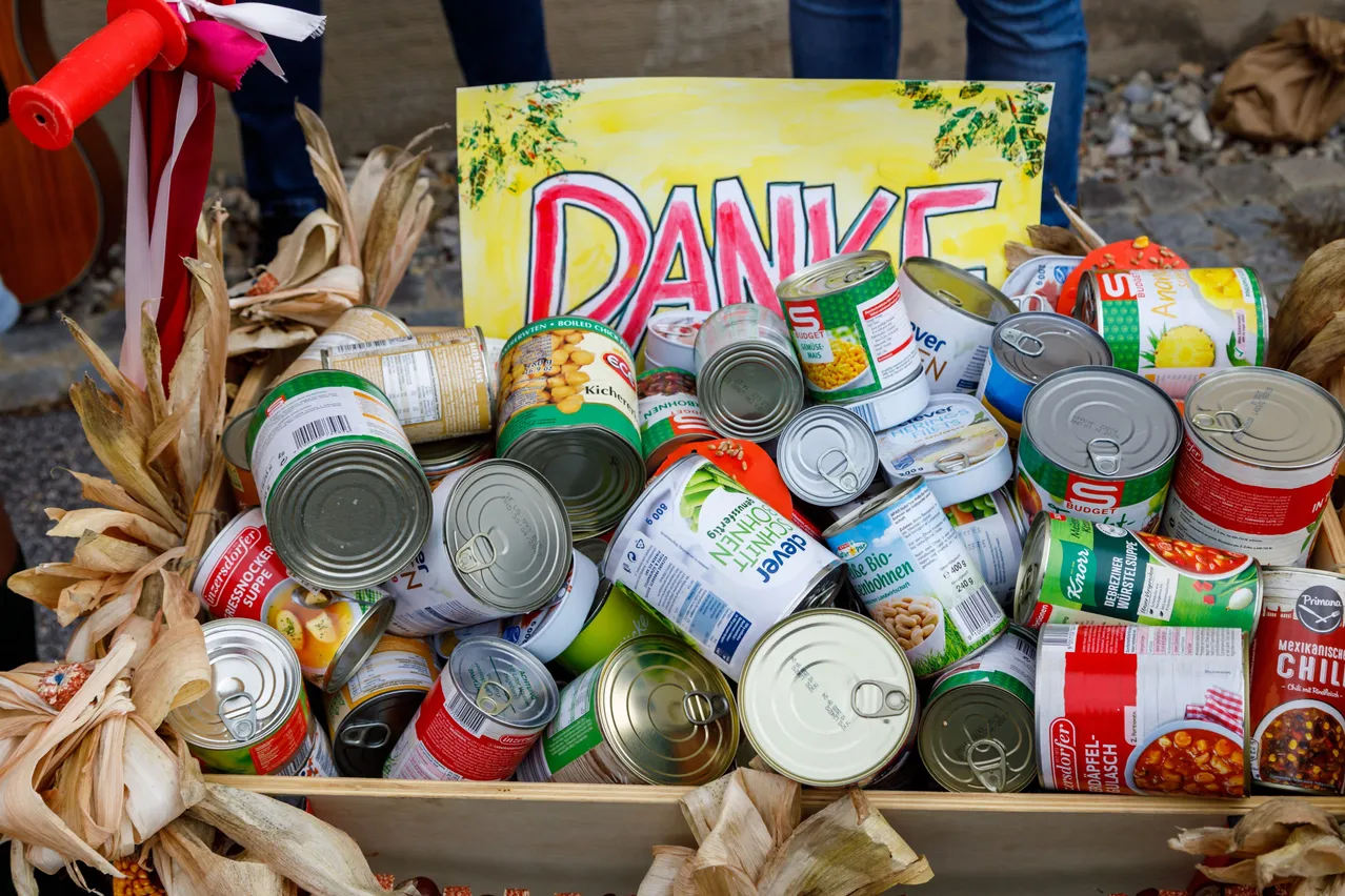 Ein hölzerner Kasten voller verschiedener Konservendosen, darunter Bohnen, Gemüse und Suppen, mit einem gelben Schild, das oben 'DANKE' steht.