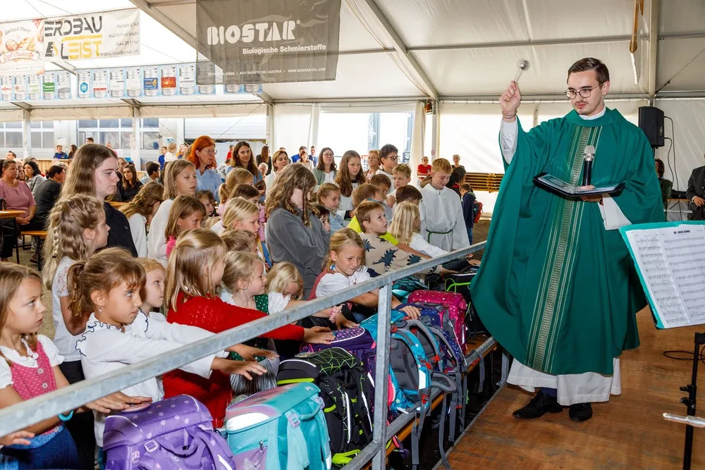 Ein Priester hält eine Glocke und ein Buch in der Hand, während er eine Gruppe von Kindern in einem Zelt anspricht. Die Kinder sind um einen Tisch mit Rucksäcken versammelt.