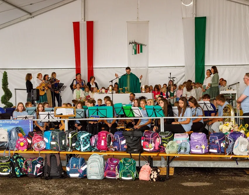 Eine Gruppe von Kindern und Erwachsenen versammelt sich in einem Zelt für eine Aufführung. Musikinstrumente sind auf einer Bühne aufgebaut, mit einem Priester in der Mitte. Taschen sind auf einer Bank vor der Bühne angeordnet.