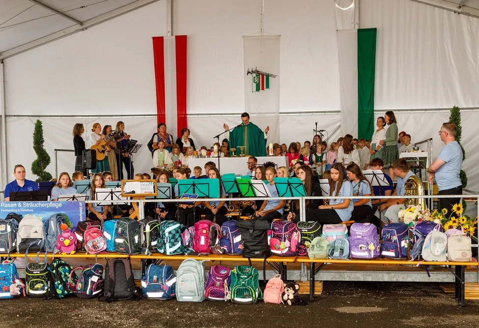 Eine Gruppe von Kindern und Erwachsenen versammelt sich in einem Zelt für eine Aufführung. Musikinstrumente sind auf einer Bühne aufgebaut, mit einem Priester in der Mitte. Taschen sind auf einer Bank vor der Bühne angeordnet.