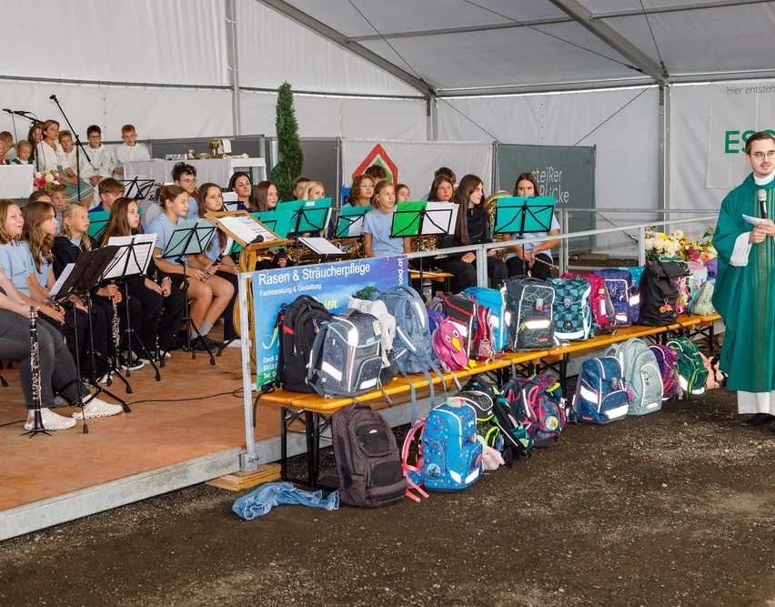 Eine Gruppe junger Musiker, wahrscheinlich Schüler, sitzt auf einer Bühne mit Musikinstrumenten. Sie werden von einem Lehrer oder Dirigenten begleitet. Vor ihnen steht eine Reihe bunter Rucksäcke, die ordentlich auf einer Bank angeordnet sind. Die Umgebung scheint ein Schulereignis oder eine Musikaufführung zu sein.