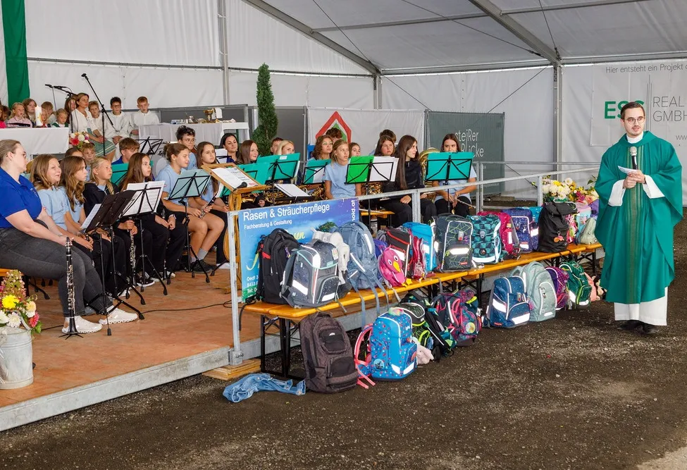 Eine Gruppe junger Musiker, wahrscheinlich Schüler, sitzt auf einer Bühne mit Musikinstrumenten. Sie werden von einem Lehrer oder Dirigenten begleitet. Vor ihnen steht eine Reihe bunter Rucksäcke, die ordentlich auf einer Bank angeordnet sind. Die Umgebung scheint ein Schulereignis oder eine Musikaufführung zu sein.