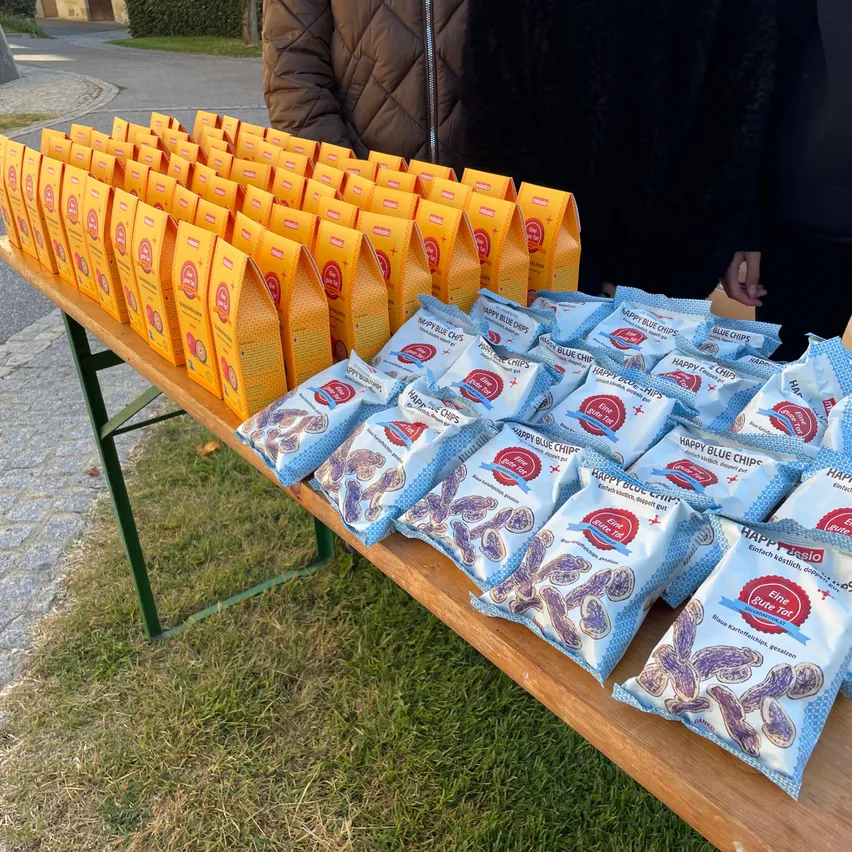 Ein Holztisch gefüllt mit mehreren Packungen Happy Blue Chips, einem Snack, im Freien ausgestellt.