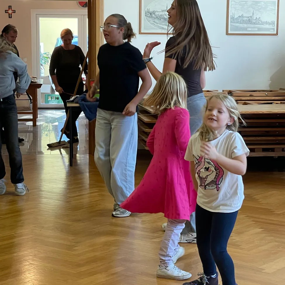 Eine Gruppe von Menschen tanzt in einem Raum mit Holzböden. Eine Frau in einem schwarzen Shirt spricht mit einem Mädchen in einem rosa Kleid.