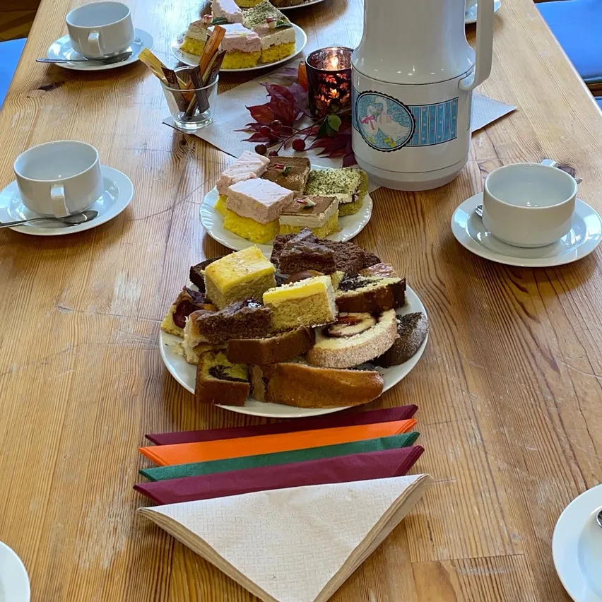 Ein Holztisch ist mit einer Vielzahl von Desserts, Kaffeetassen, Untertassen und einem Teekessel besetzt. Der Tisch ist mit bunten Servietten und einer Kerze geschmückt.