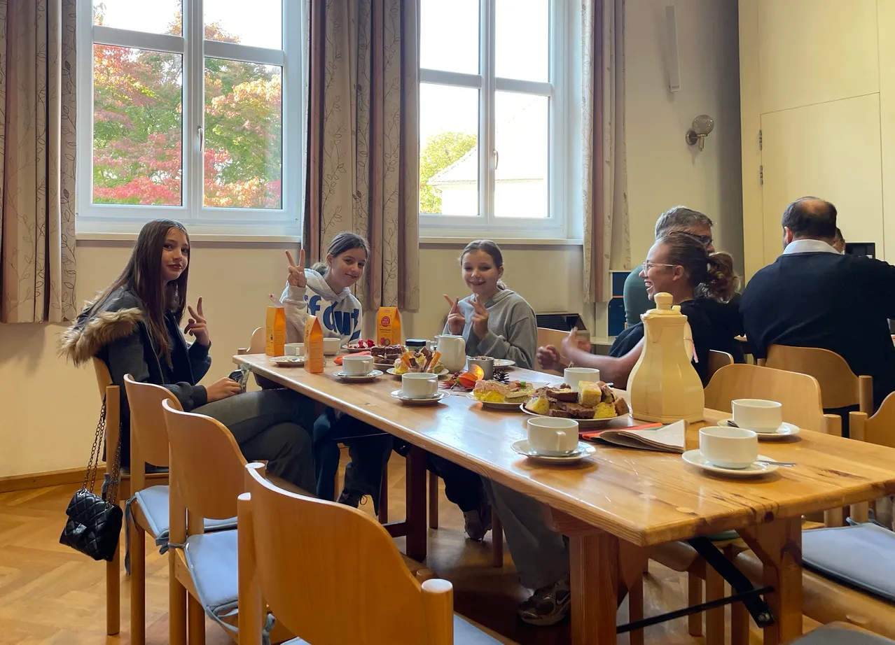 Eine Gruppe von Menschen sitzt um einen Tisch mit Tassen, Untertassen und verschiedenen Speisen. Der Tisch ist mit Tellern, Tassen und Untertassen besetzt. Der Raum hat große Fenster mit Vorhängen.